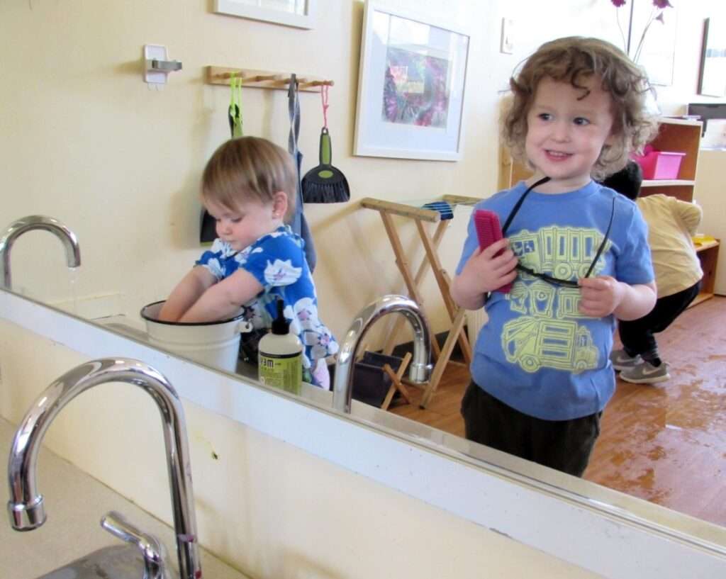 Lyonsgate Montessori Toddler student admiring the view in the mirror while friends engage in water work.