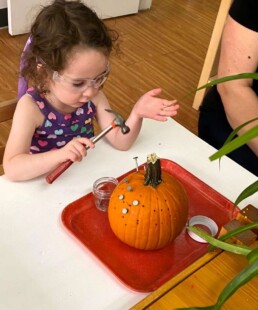Lyonsgate Montessori School Casa student developing hand-eye coordination and by hammering nails into a pumpkin.