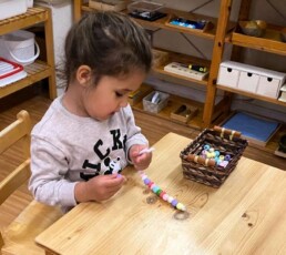 Lyonsgate Montessori School Casa student developing fine motor skills, concentration, and creativity with a bead-threading activity.