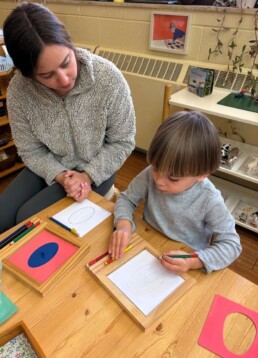 Lyonsgate Montessori School Casa student receiving a presentation of the iconic Montessori Metal Insets material that develops dexterity and control, introduces geometry vocabulary, and is an artistic activity.