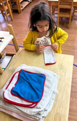 Lyonsgate Montessori School Casa student folding the classroom cloths help care for her classroom community while building motor skills for precision, and concentration.